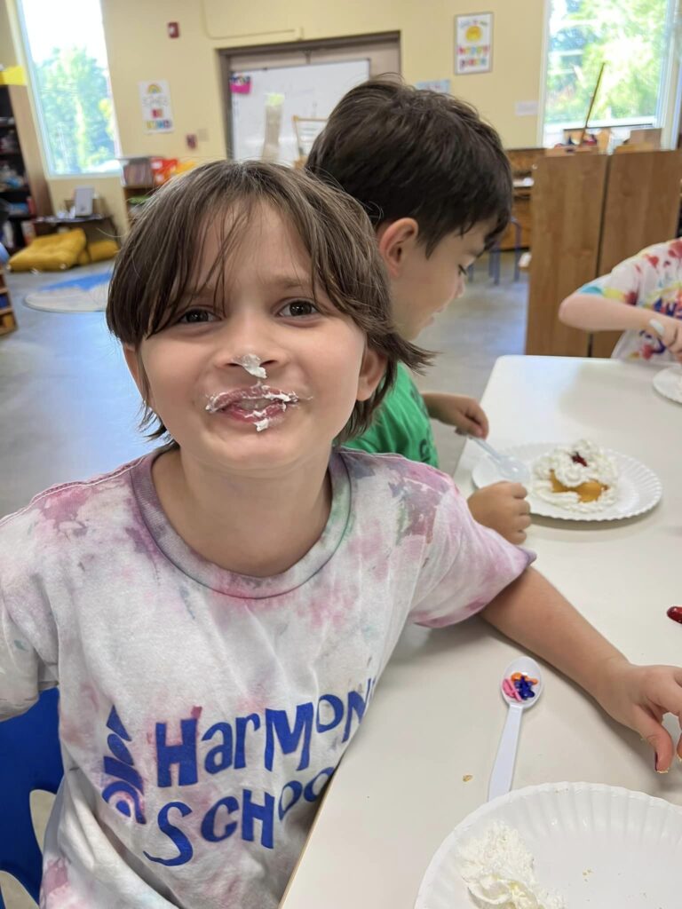 A child with light skin and short brown hair smiling at a shared table wearing a Harmony School shirt and with icing on their nose and mouth.