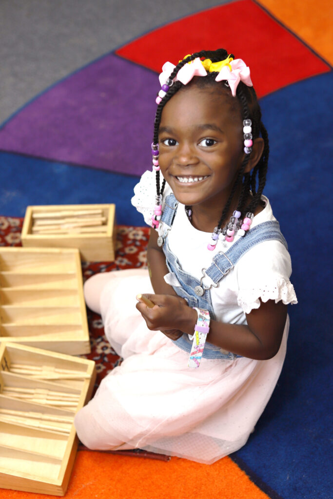 A girl with brown skin and braids sits on a colorful carpeted floor using Montessori math materials to learn counting.