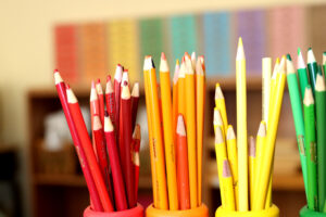 Colorful pencils holders displaying sorted red, orange, yellow, and green pencils against a blurred colorful classroom.