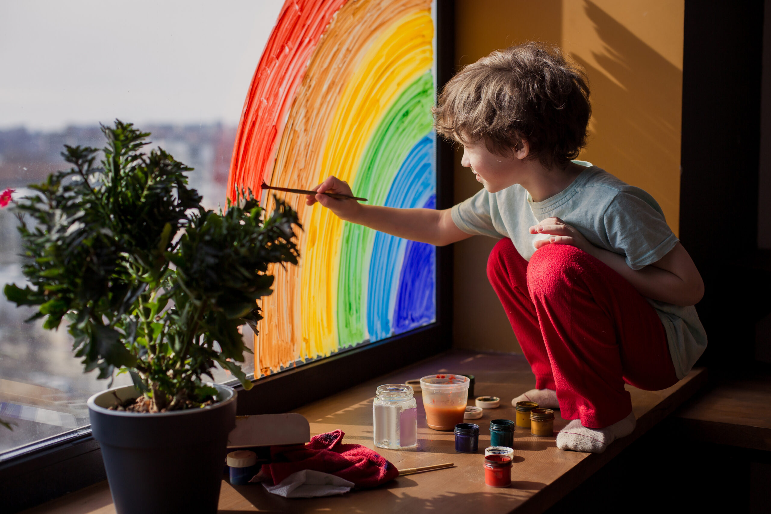 A child with light skin stopped on a window seat painting a rainbow onto the corner of a large window overlooking a blurred city. There are paints laying around him and a potted plant.
