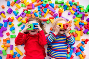 Two children with light skin laying on a floor surrounded by colorful blocks and holding them over blocks over their eyes like goggles.