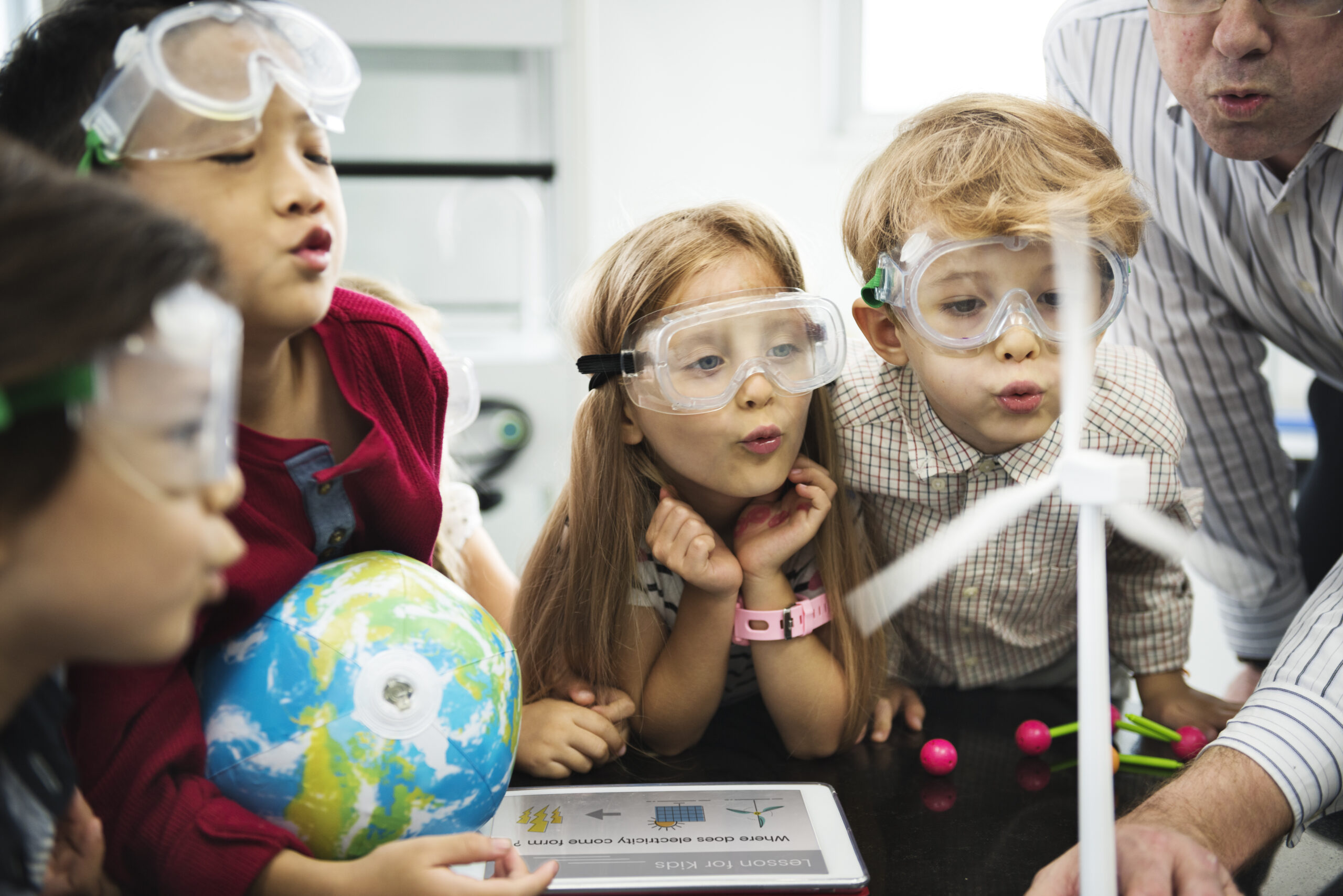 A diverse group of children wearing safety goggles and blowing onto a handmade windmill with other items from a science class around them.